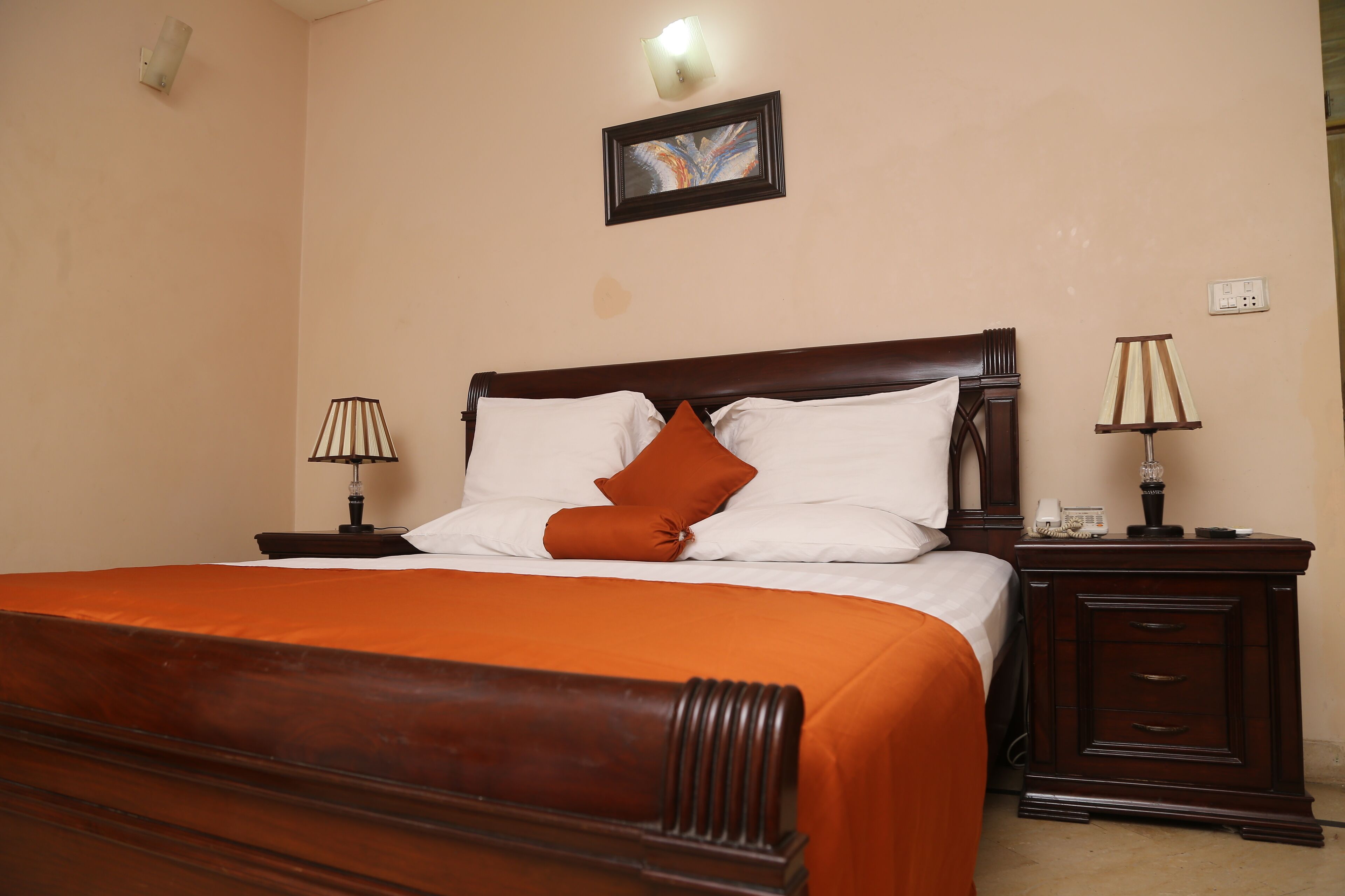 A neatly made bed with an orange bedspread and two pillows, a wooden nightstand with a lamp, and a wall-mounted picture.