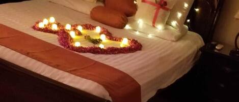 A neatly made bed with a heart-shaped arrangement of candles and rose petals on the floor.