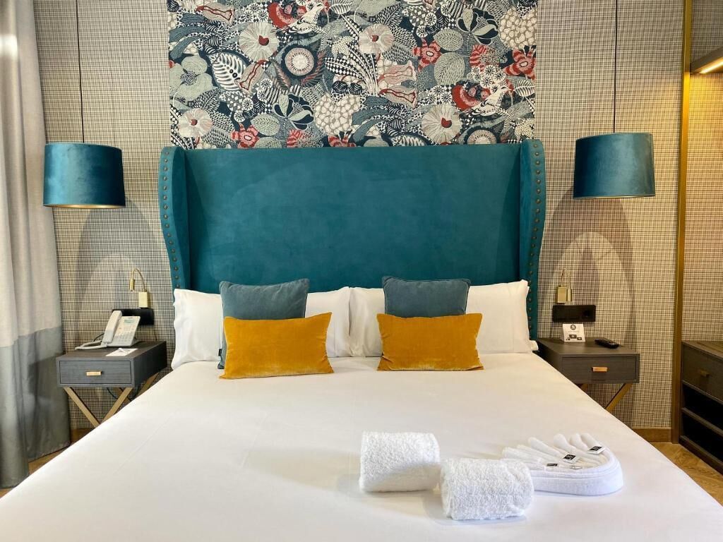Una cama bien hecha con dos almohadas, una mesita de noche con lámpara y un papel tapiz estampado de fondo.