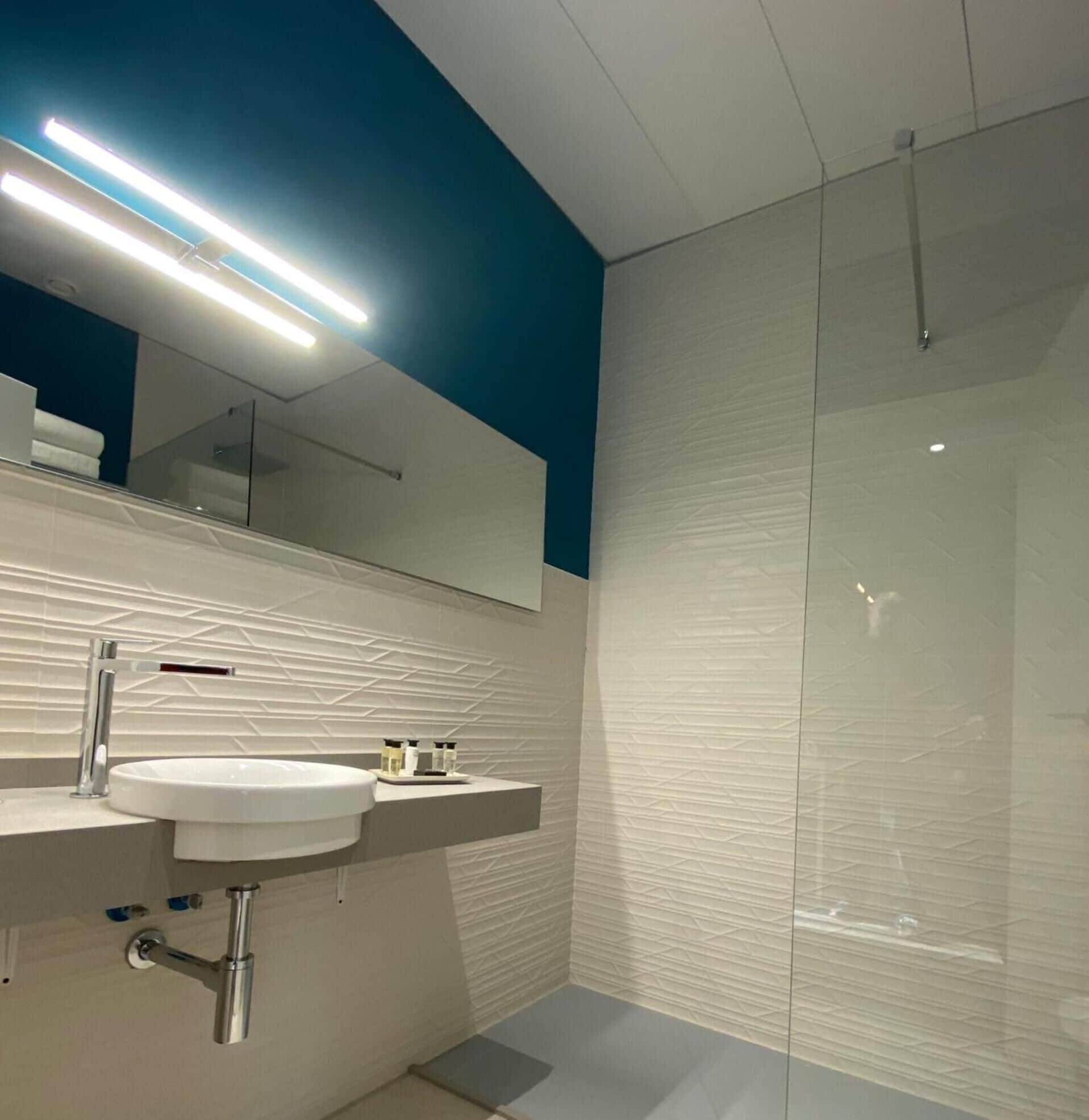 Un baño moderno con lavamanos blanco, espejo y cabina de ducha con mampara de vidrio.