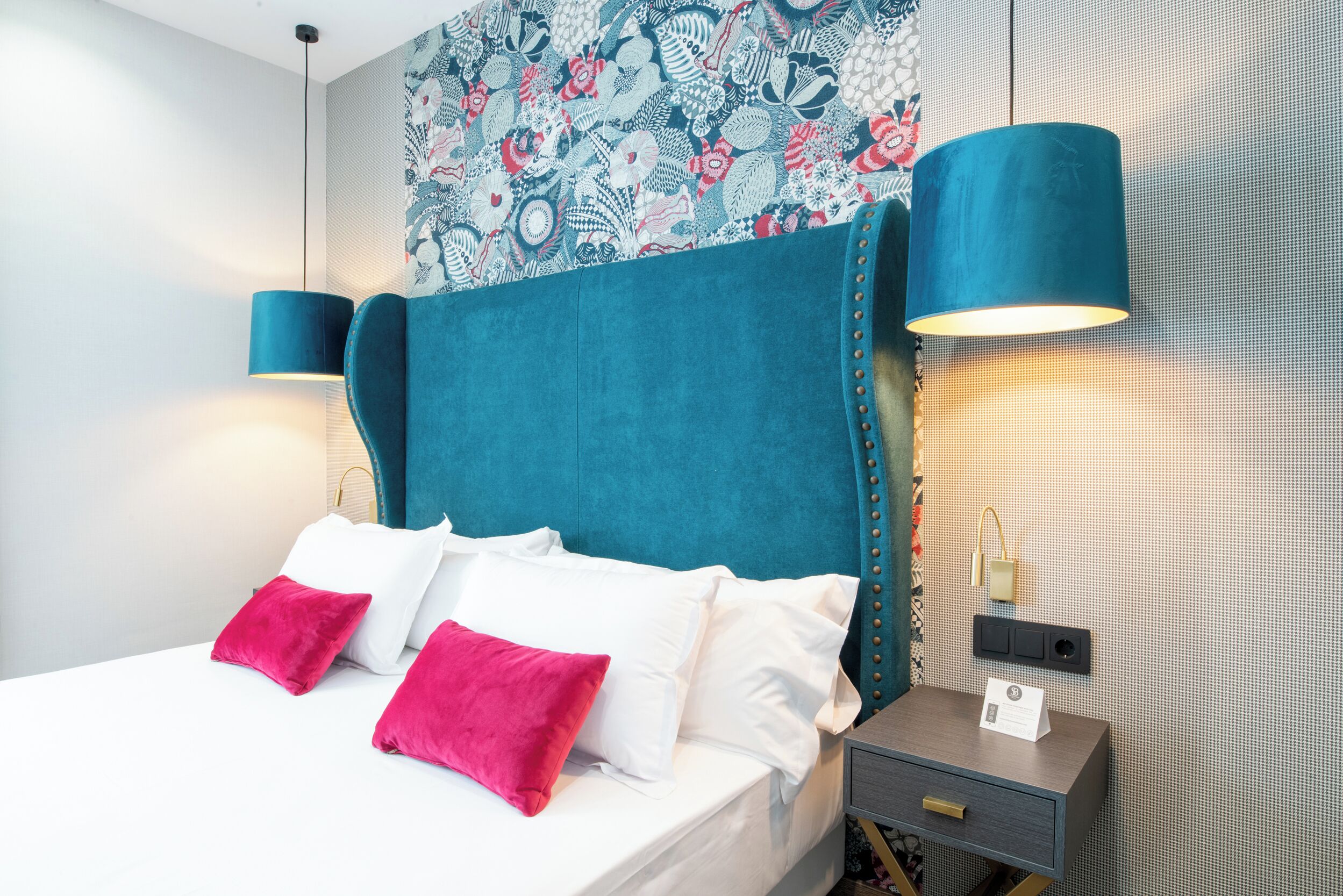 Una habitación de hotel moderna con una cama grande, almohadas blancas y rosas, cabecera color aguamarina y dos lámparas fijadas a la pared.