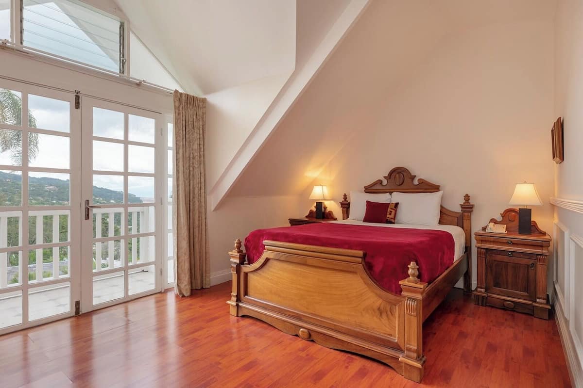 A bedroom with a wooden bed, two bedside tables, a large window with a view, and a balcony.