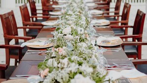 A long dining table with chairs, set for a meal, surrounded by a white fence and palm trees in the background. - Fogo Resort (San Antonio)