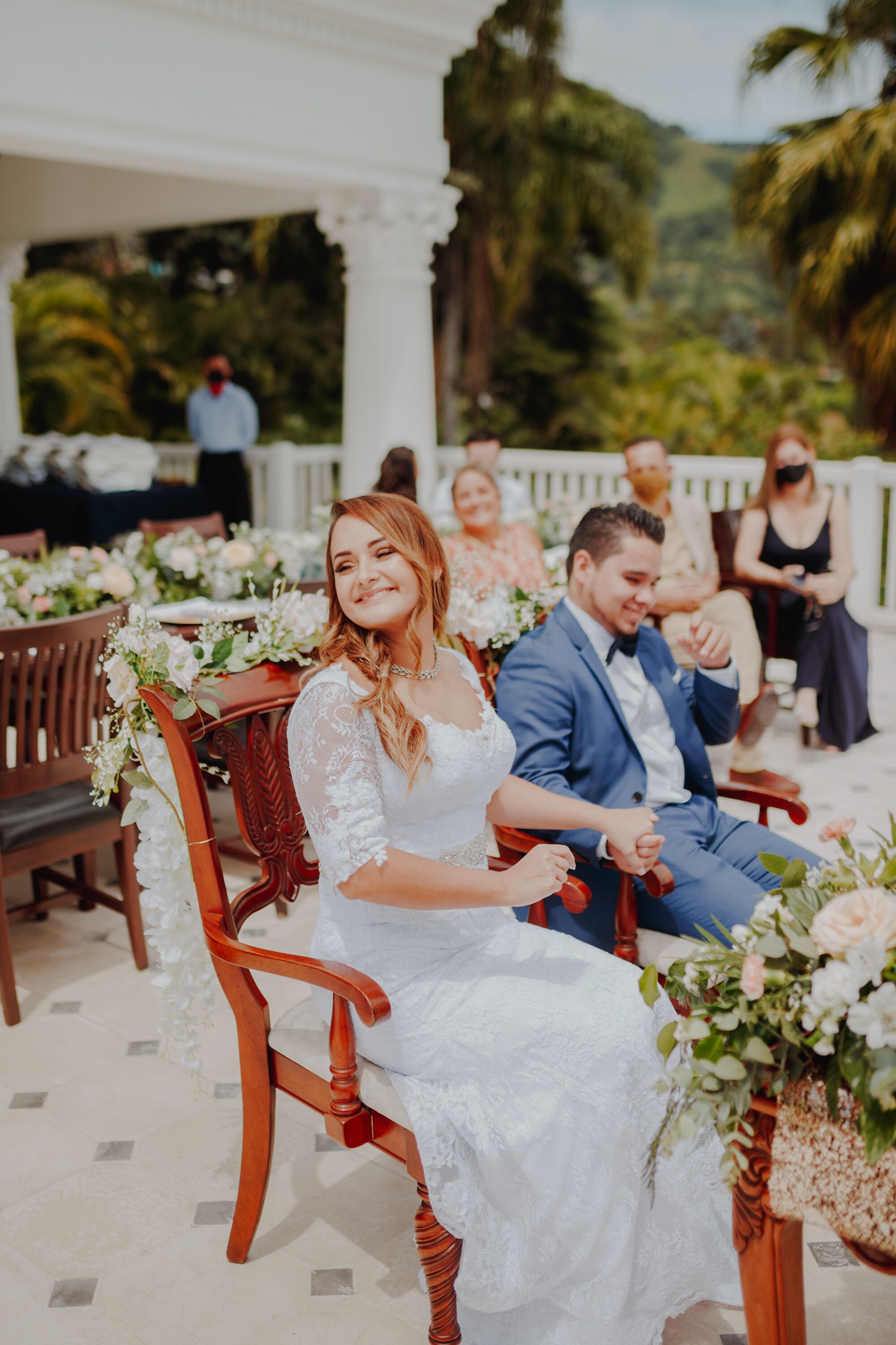 A wedding ceremony with a bride and groom seated on wooden chairs adorned with floral arrangements.