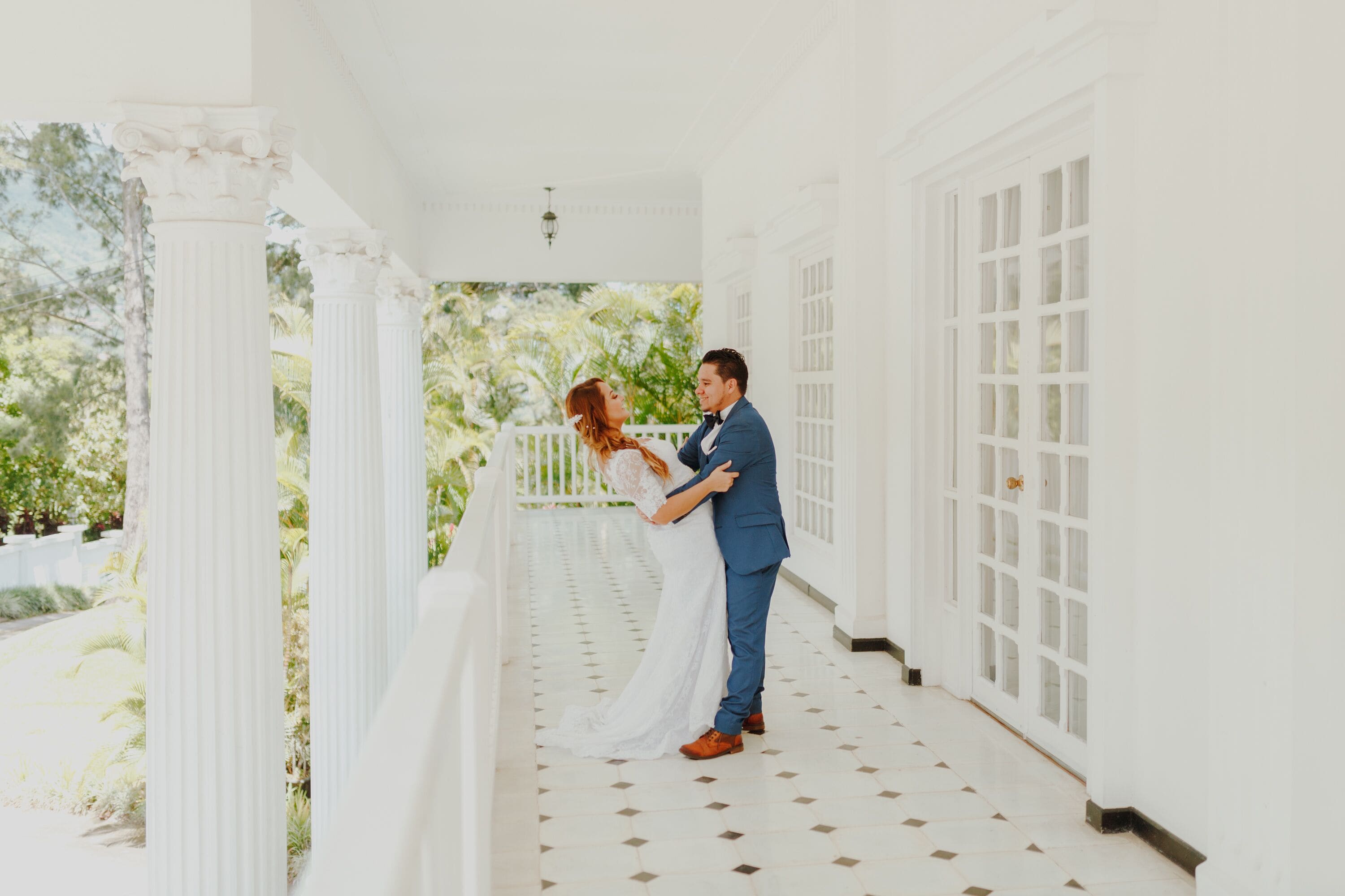 A couple embracing on a white porch with columns and a view of greenery.