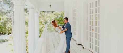 A couple embracing on a white porch with columns and a view of greenery.