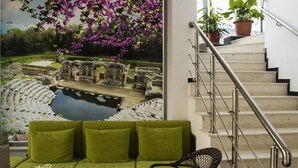 A green sofa and a wicker chair in a room with a mural of a garden and ruins. - Hotel Titania (Sarandë)