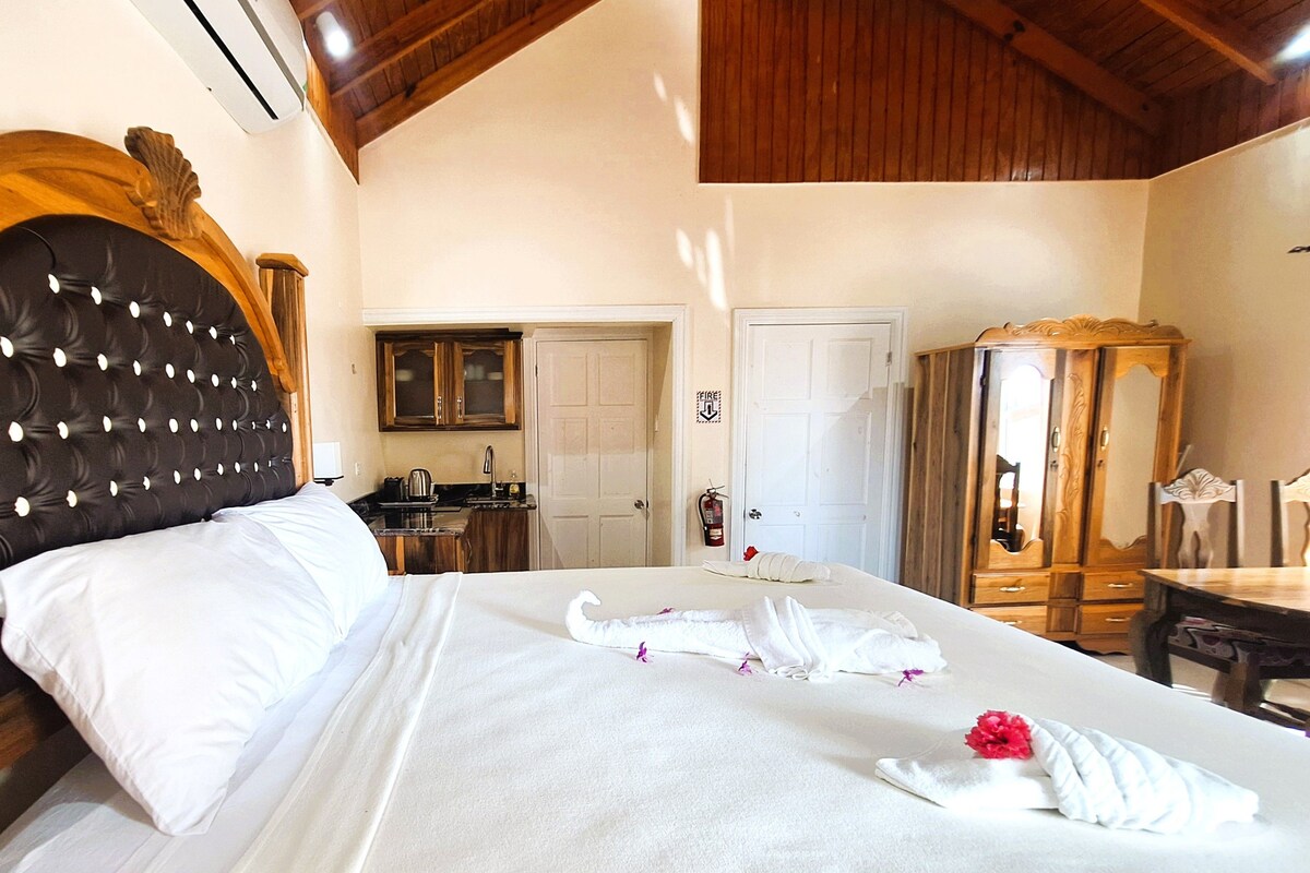 A hotel room with a large bed, a wooden headboard, a wooden wardrobe, a small table, and a kitchenette in the background.
