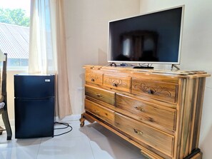Een houten dressoir met laden en een flatscreen televisie erop, een kleine zwarte koelkast, en een raam met gordijnen.