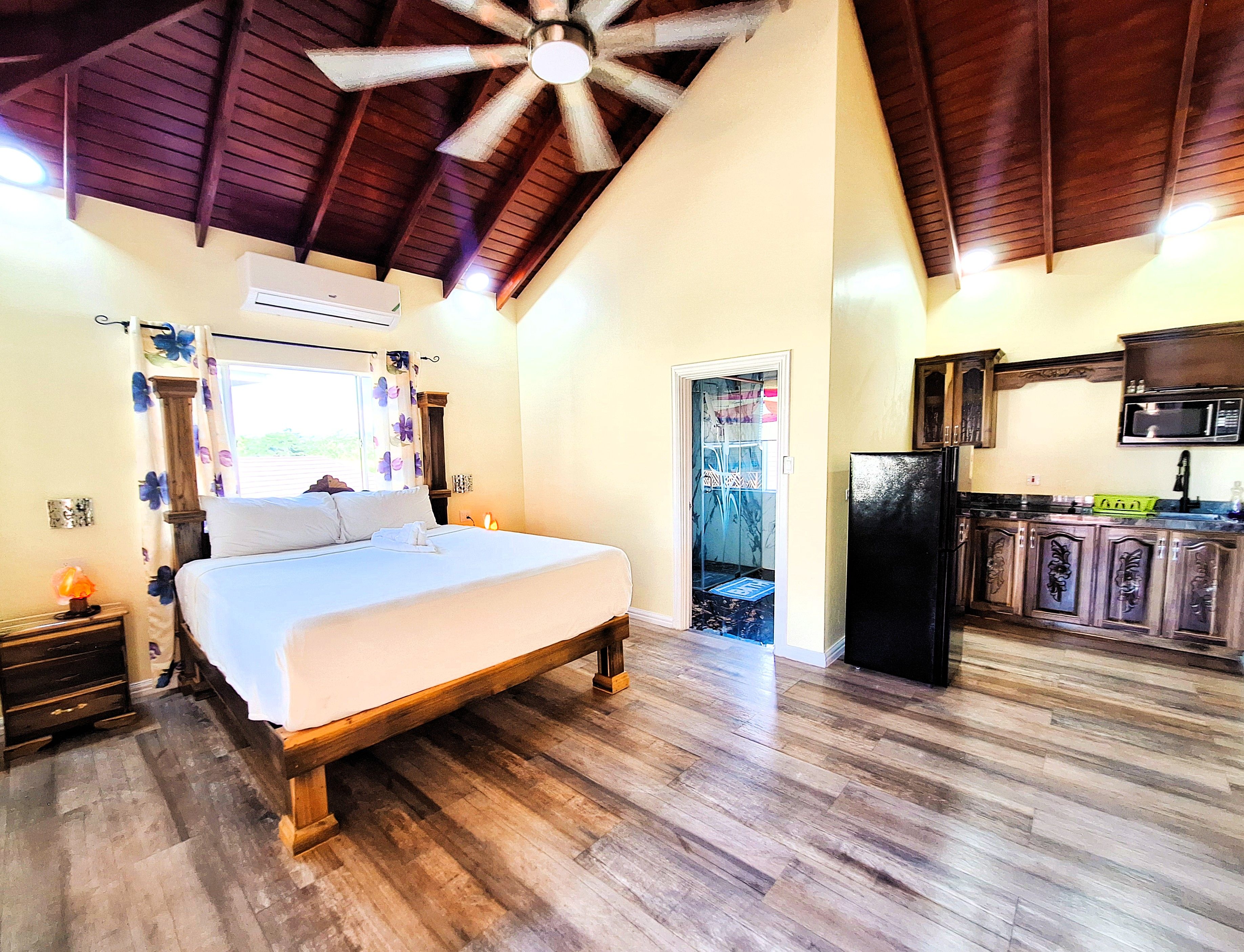 A bedroom with a wooden bed, a ceiling fan, and a view of a kitchen with a refrigerator and microwave.