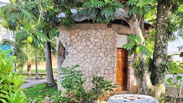 Un edificio de piedra con puerta de madera, rodeado de abundante vegetación y una mesa de mosaicos.