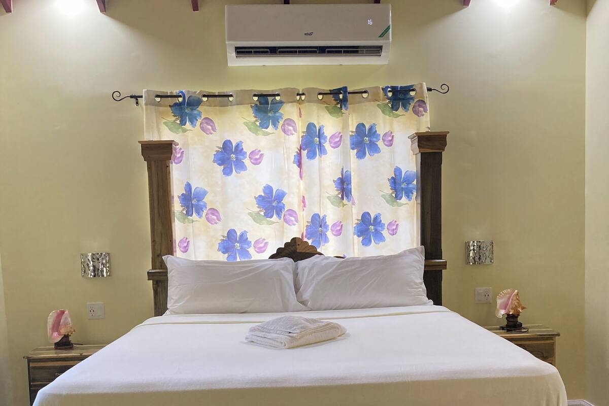 A bedroom with a wooden bed frame, white bedding, a floral curtain, an air conditioning unit, and a bedside table with a lamp.