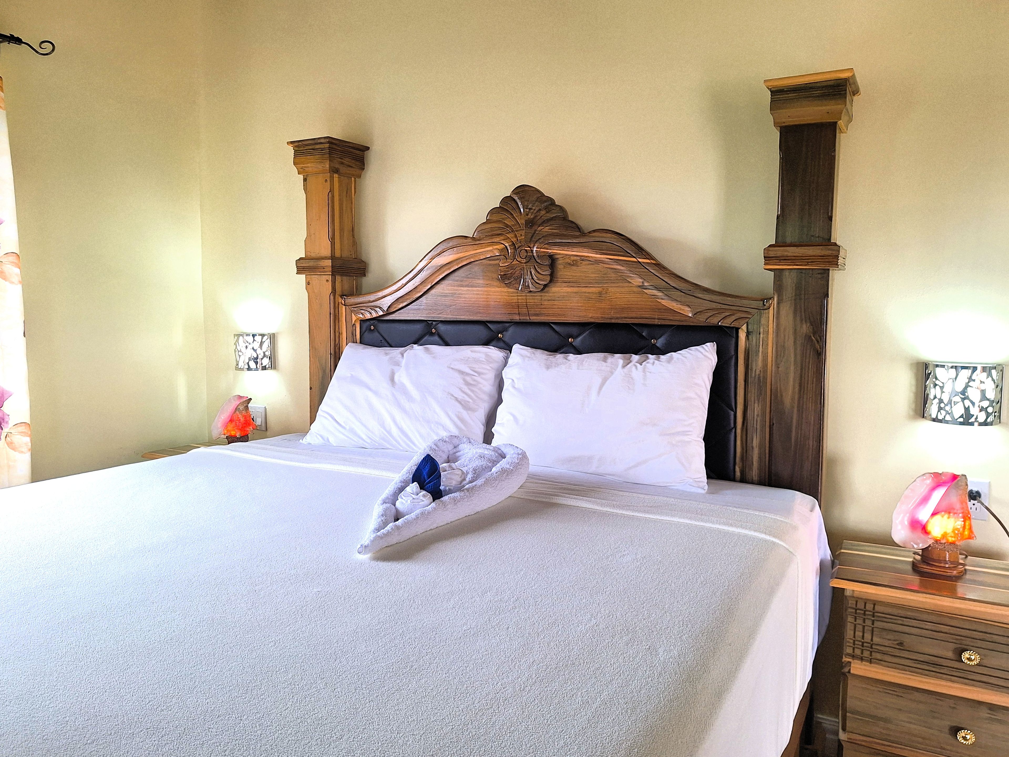 Ein ordentlich bezogenes Bett mit weißer Daunendecke, einem Holz-Kopfstück und einem Nachttisch mit Lampe.