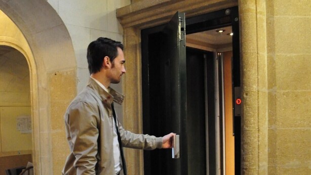 Un hombre con una maleta de pie junto a la puerta abierta de un ascensor en un edificio de arquitectura de piedra.