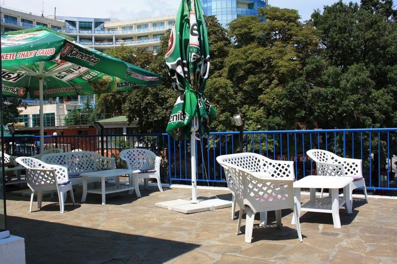 Outdoor seating with white chairs and a green umbrella, a blue metal fence, and trees in the background.