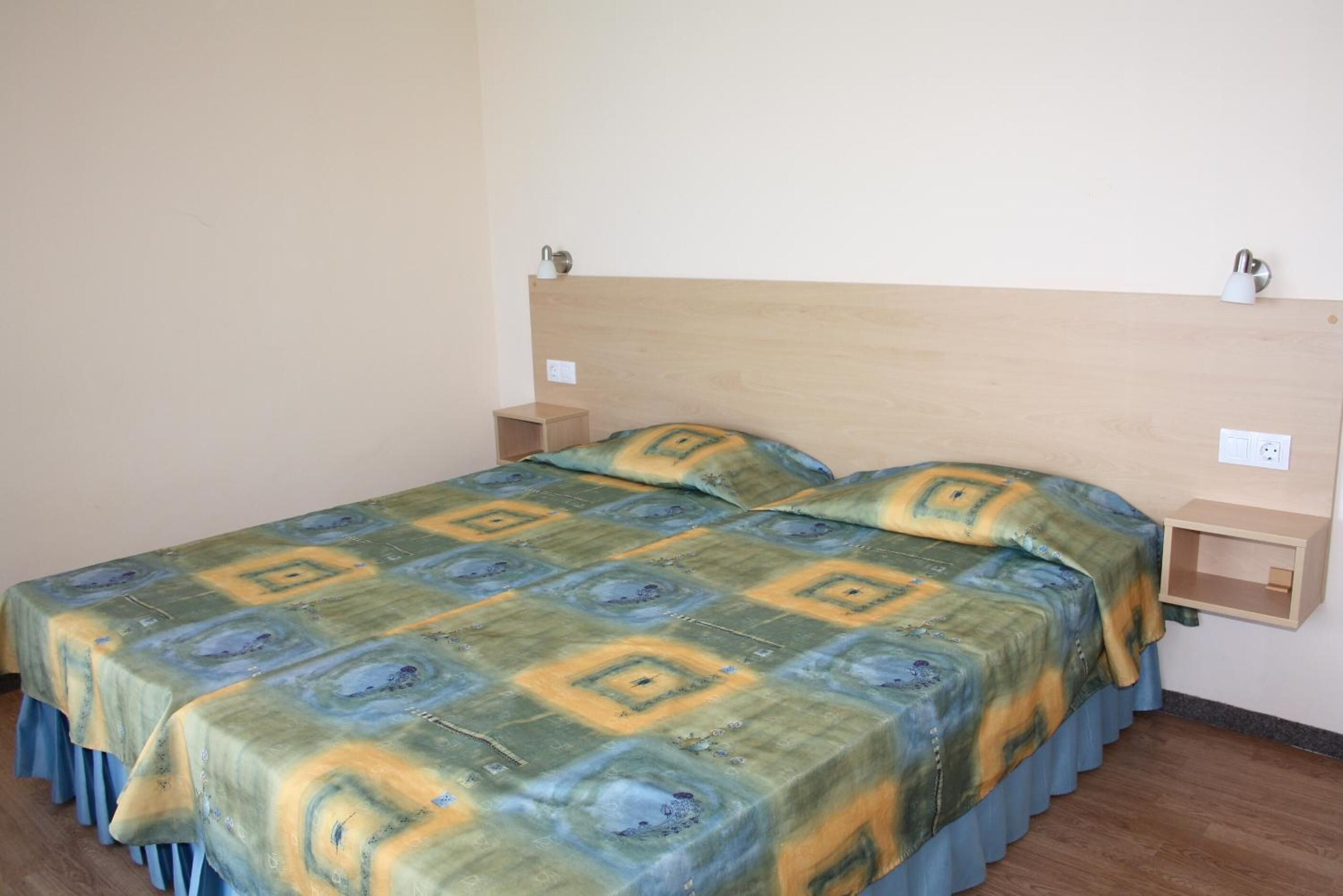 A single bed with a patterned bedspread, a wooden headboard, bedside tables, and a wall socket.