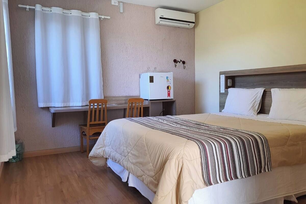 A hotel room with a bed, a desk, a chair, an air conditioner, and a wall-mounted heater.