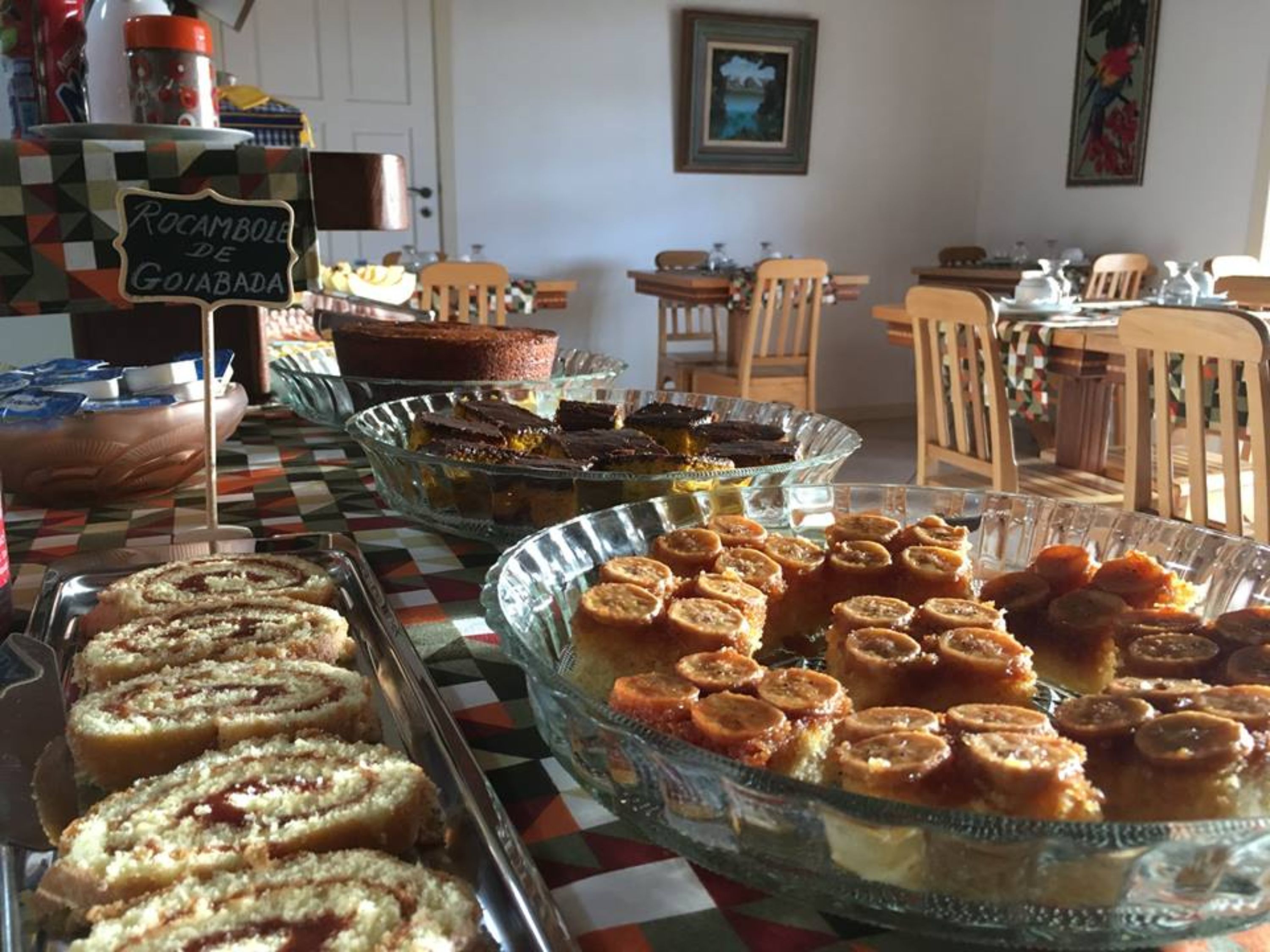 A buffet with various desserts, including cakes and pastries, in a dining area with wooden chairs and a checkered tablecloth.