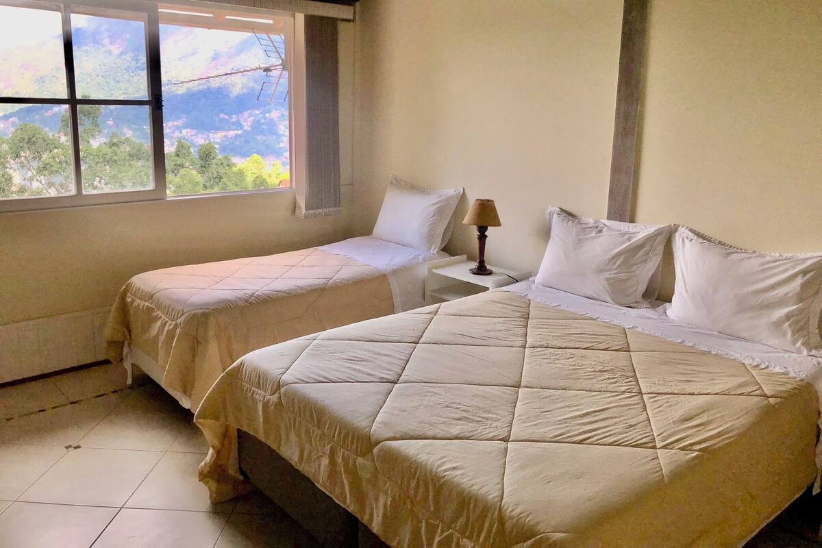 A room with two beds, a window with a view of greenery and mountains, and a small table with a lamp.