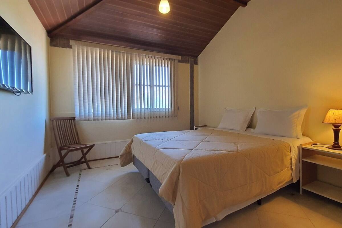 A bedroom with a bed, a chair, a nightstand with a lamp, a window with blinds, and a ceiling light.