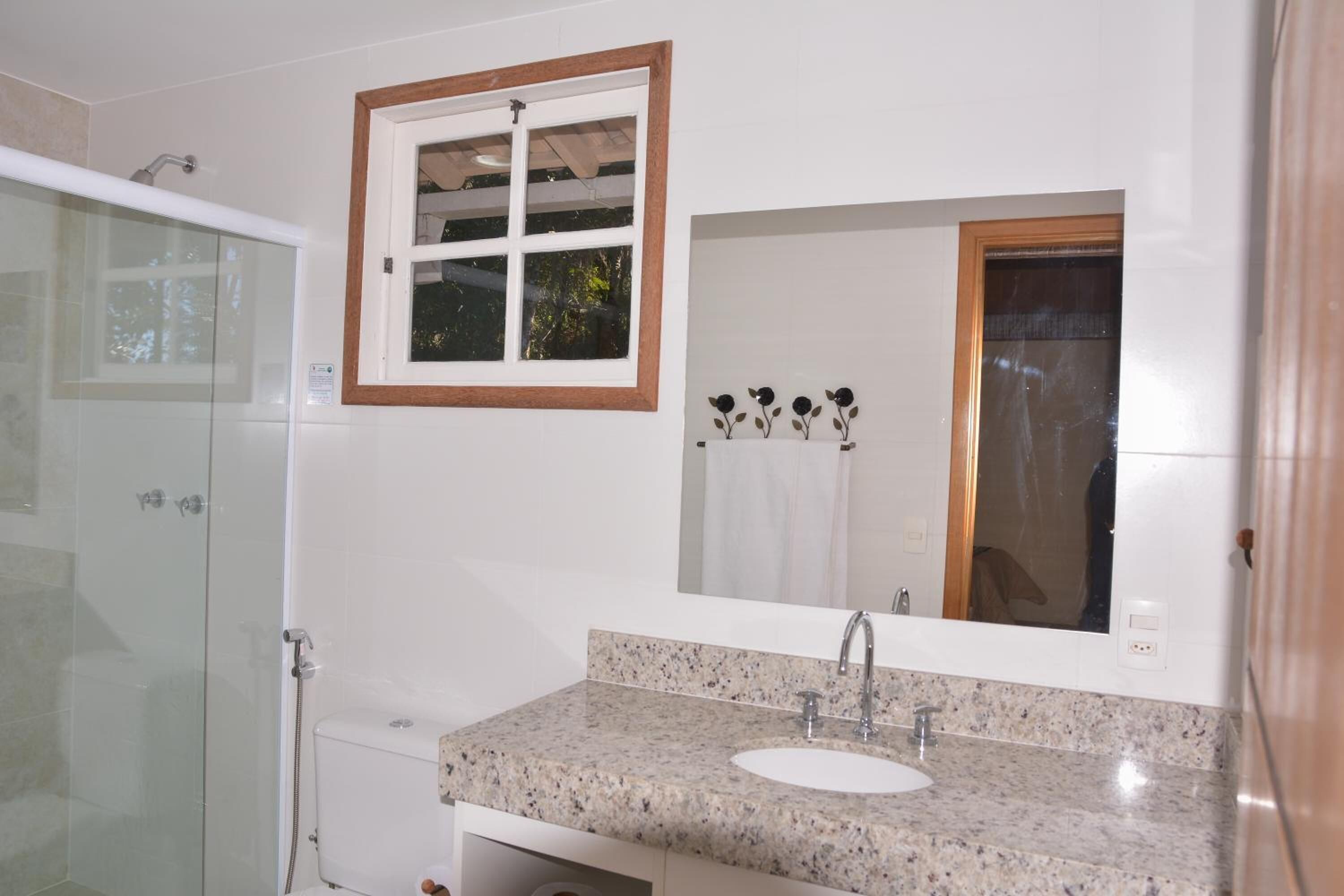 Une salle de bain dotée d’une douche à parois de verre, d’un lavabo sur un plan de travail en granit et d’un grand miroir surmonté d’une fenêtre à cadre en bois.