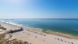 A beach with a pier extending into the ocean, a sandy beach with umbrellas, and a building with a blue roof. - San Carlos Condos (Gulf Shores)