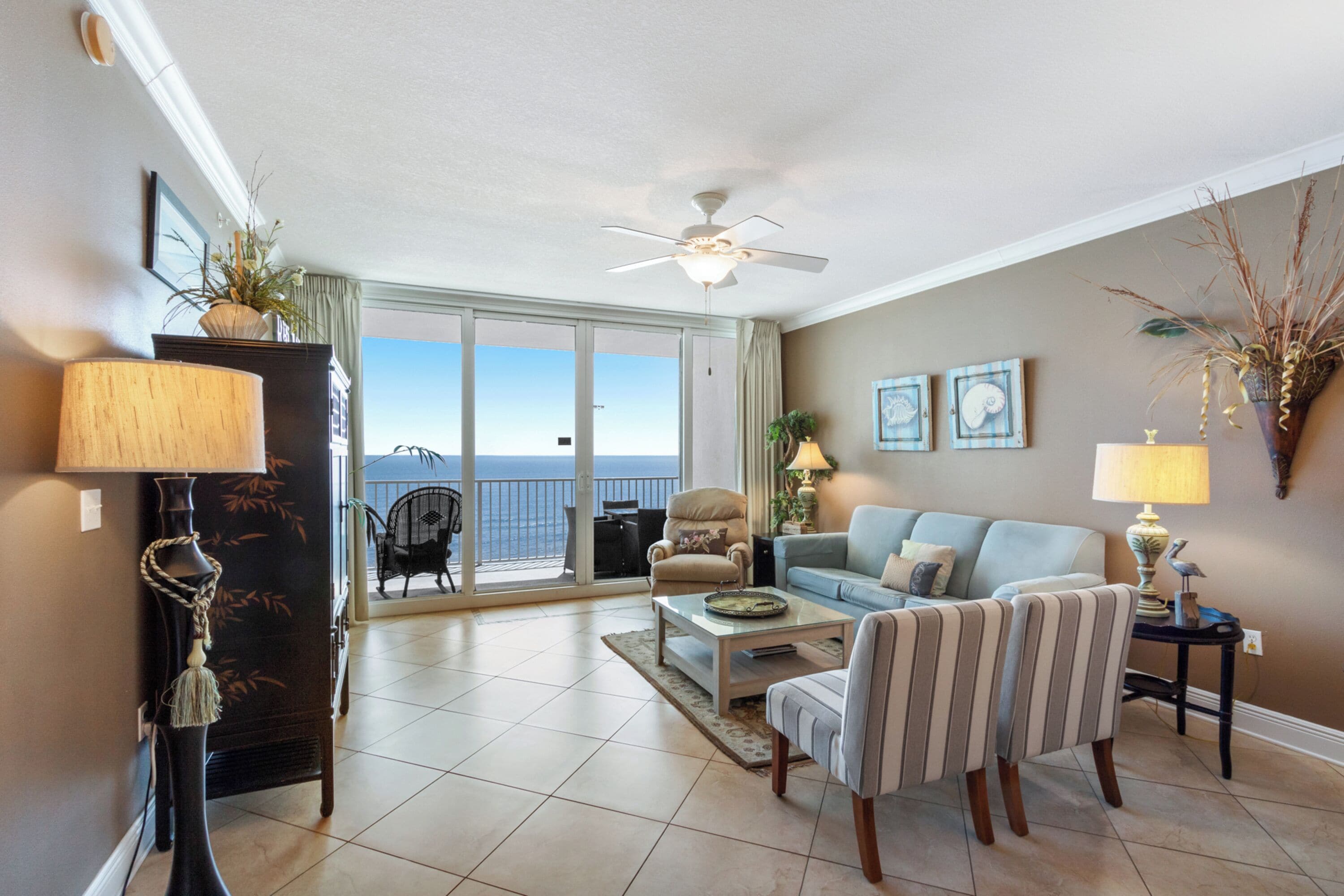 A living room with a sofa, armchairs, a coffee table, and a ceiling fan. There is a view of the ocean through large windows.