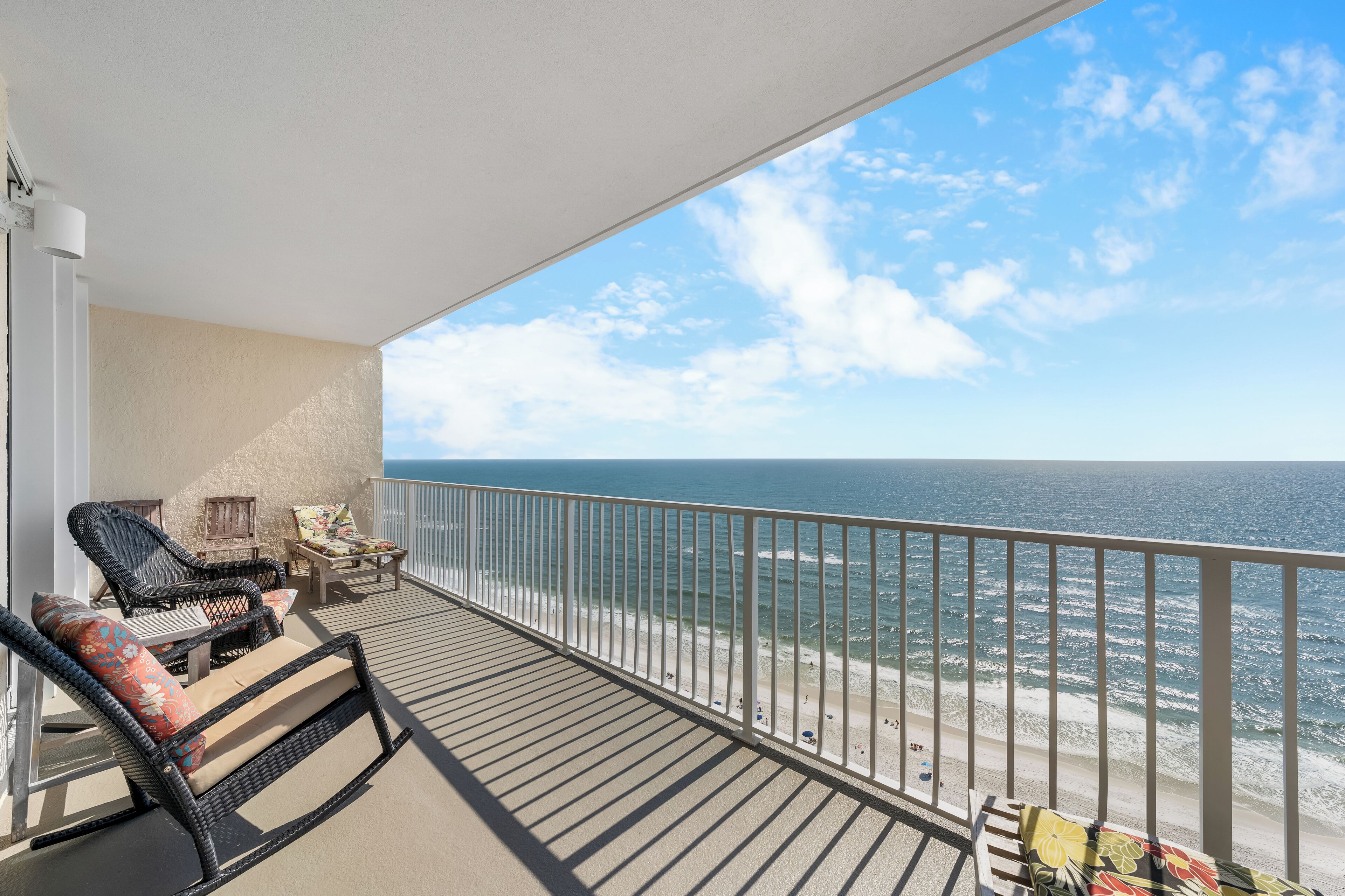 A balcony with wicker chairs and a table, overlooking a beach and ocean.