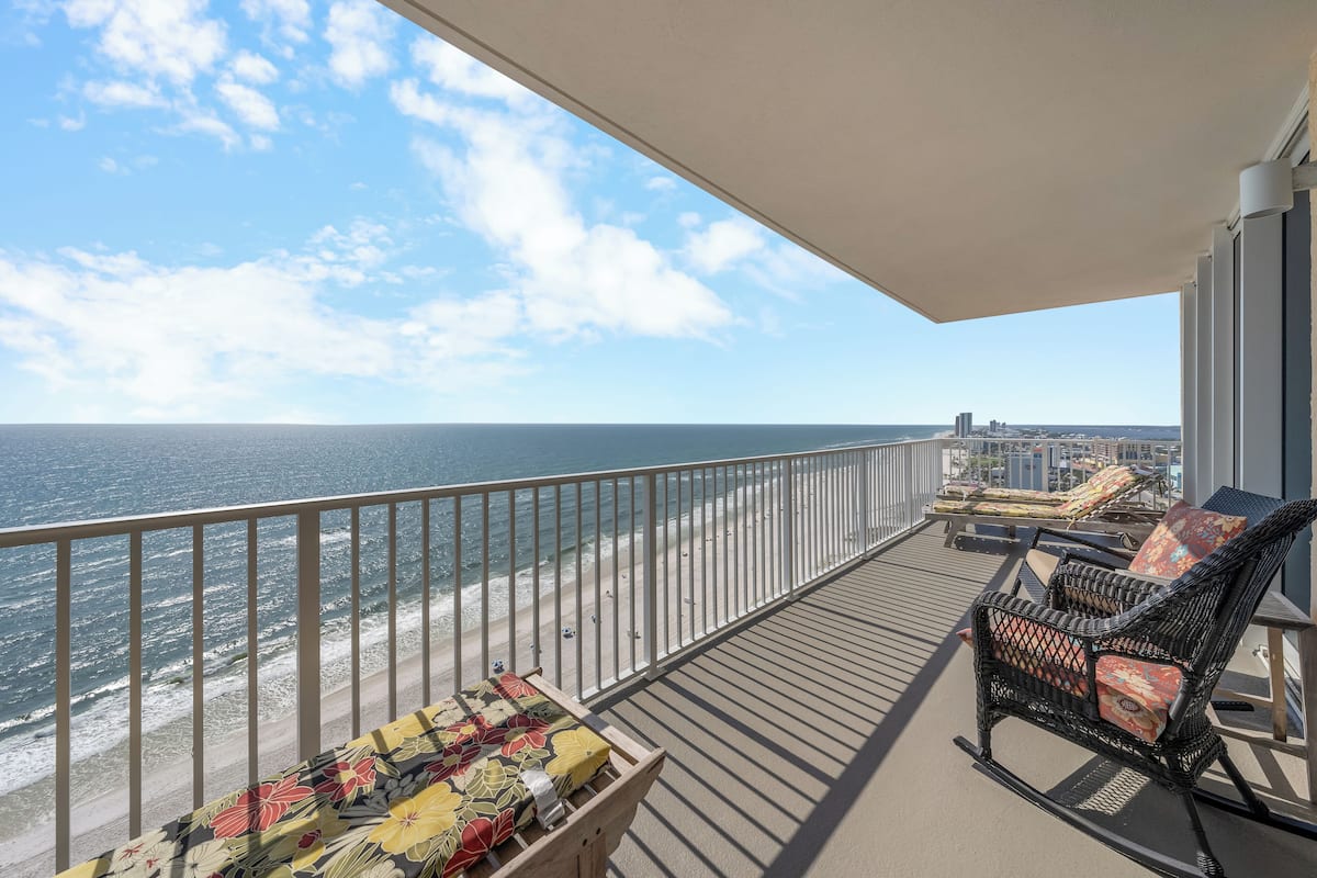 Un balcon avec vue sur la plage et l’océan, une chaise en osier et une table recouverte d’une nappe à motifs floraux.