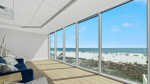 A spacious room with large windows overlooking a beach, featuring a blue sofa and a wooden floor. - San Carlos Condos (Gulf Shores)
