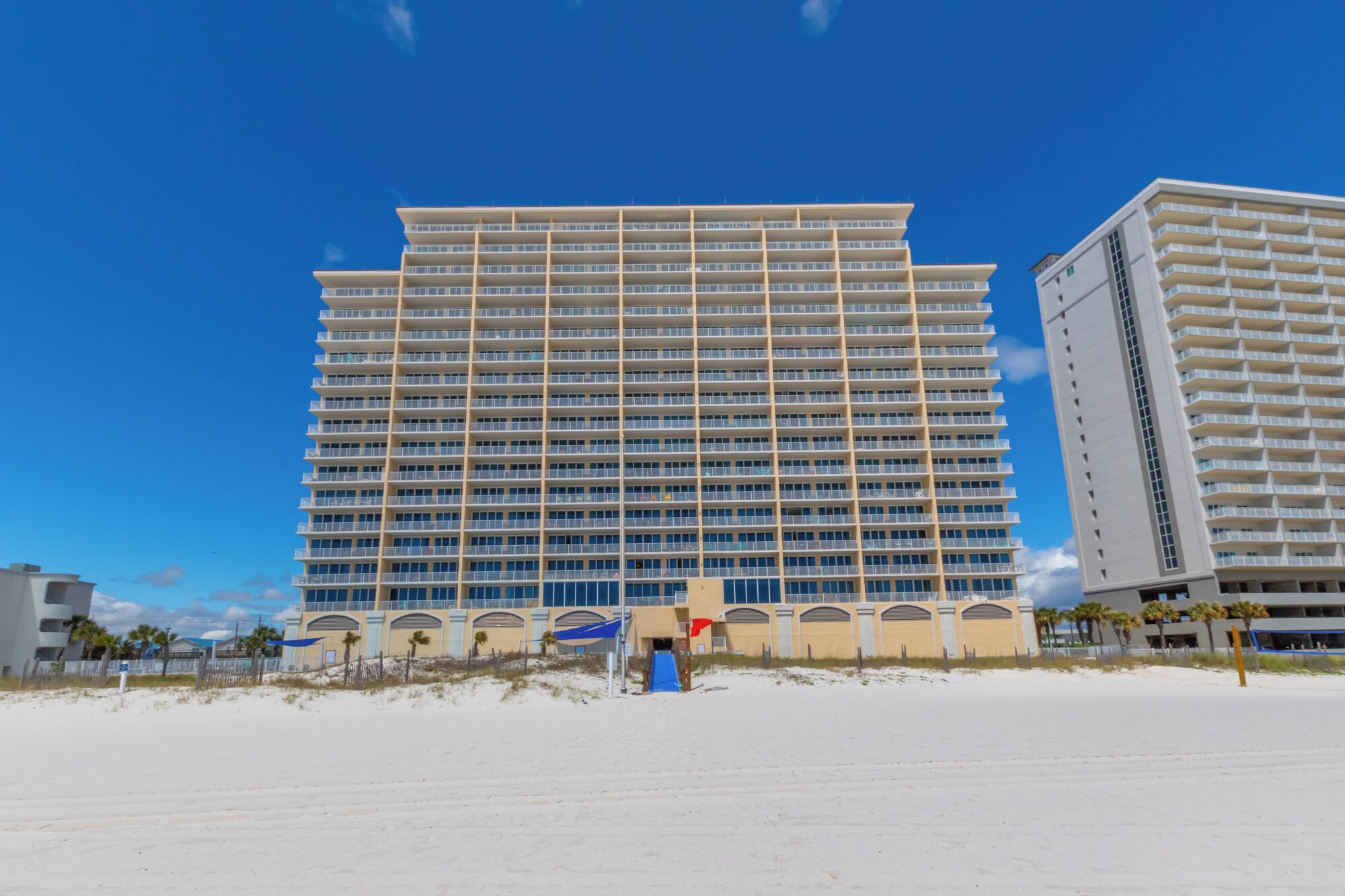 A high-rise hotel with multiple balconies, a beach in the foreground, and a clear blue sky.