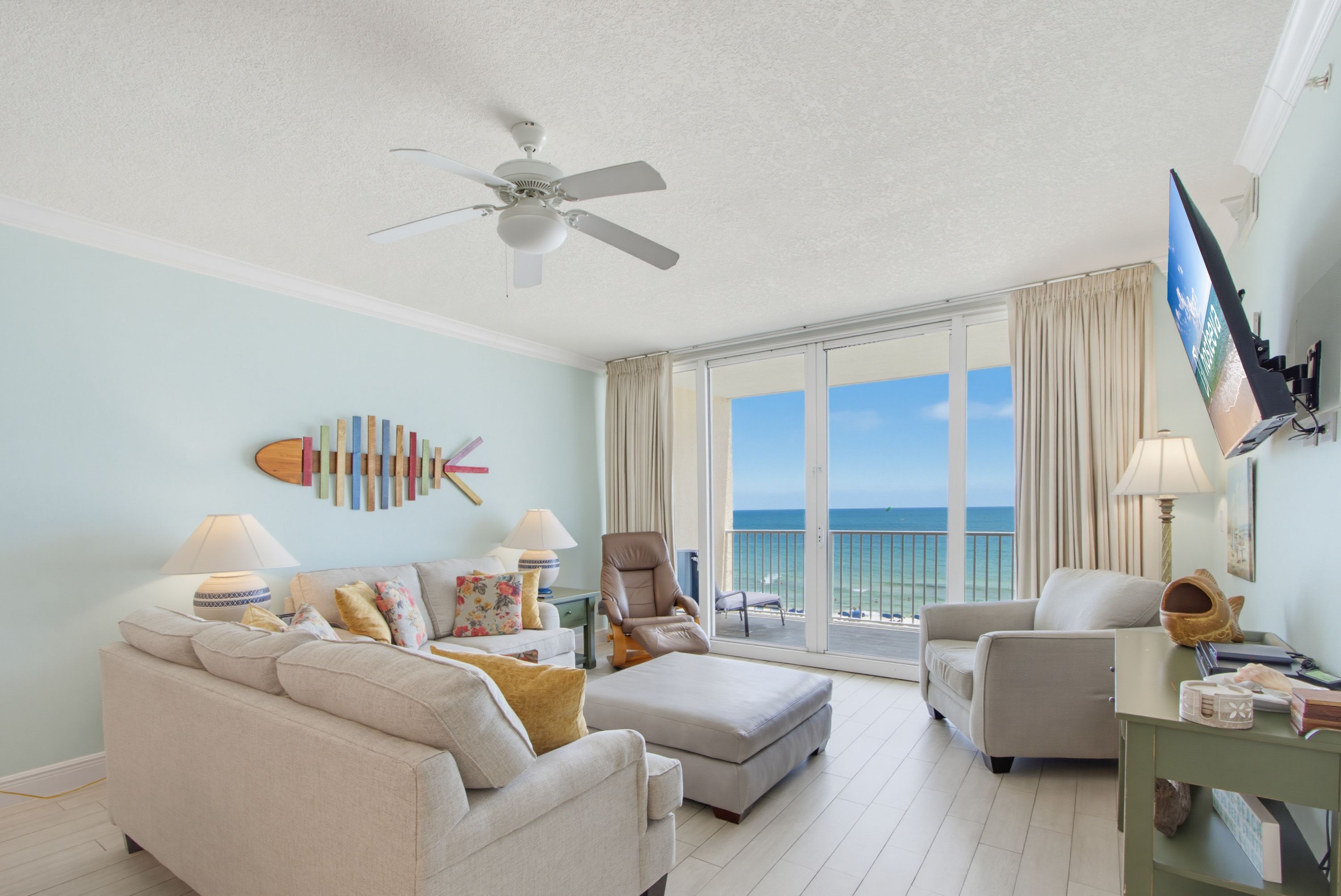 A spacious living room with a balcony, a ceiling fan, a flat-screen TV, and a view of the ocean.