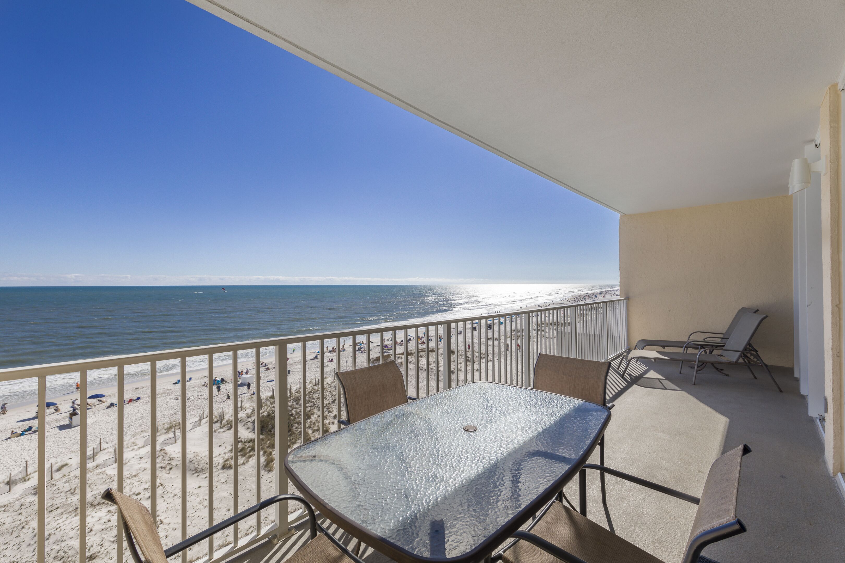Varanda com mesa e cadeiras de vidro, com vista para uma praia arenosa e o oceano.