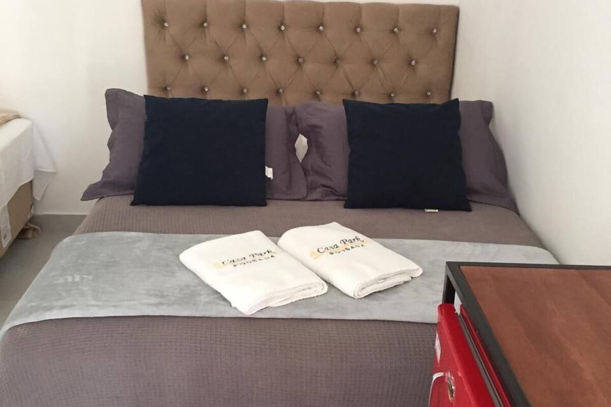 A neatly made bed with a tufted headboard, two pillows, a grey blanket, and two towels with 'Casa Park' branding.