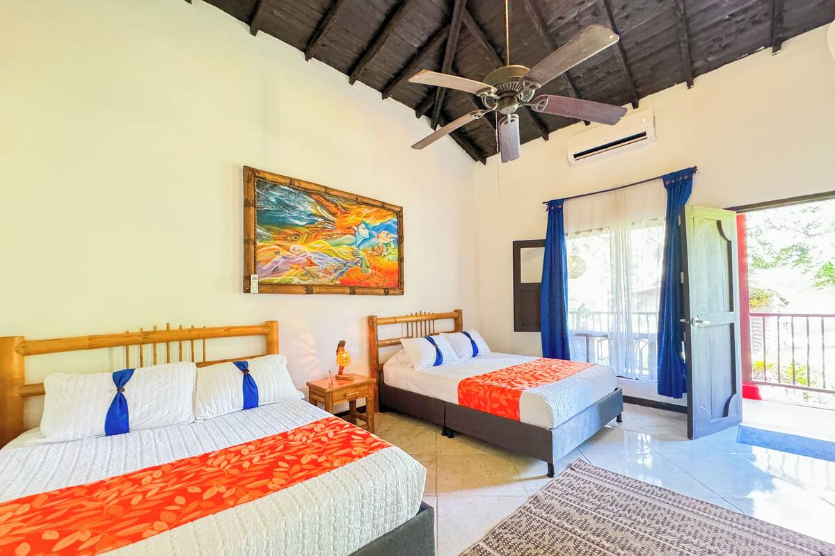 Una habitación con dos camas, ventilador de techo, un cuadro y un balcón con vistas.