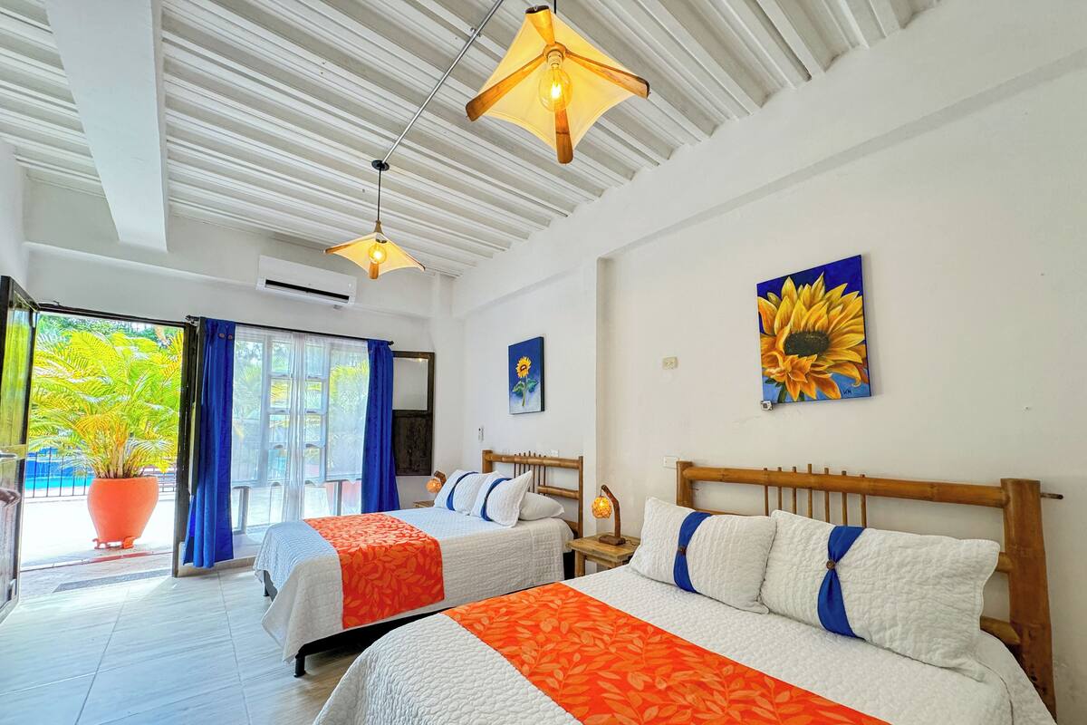 Habitación de hotel con dos camas, un ventanal con cortinas, una planta en maceta y un cuadro de un girasol.