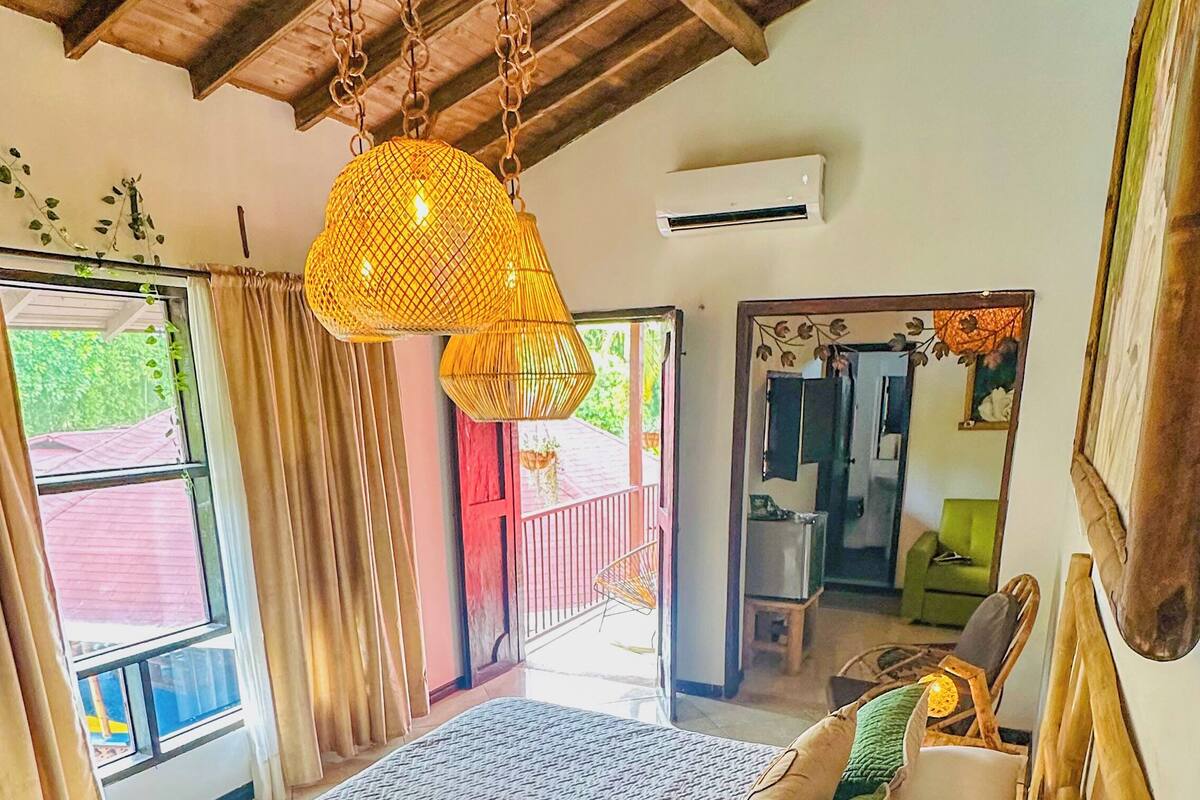 Un dormitorio con cama, lámpara colgante, ventana y puerta que da a otra habitación.