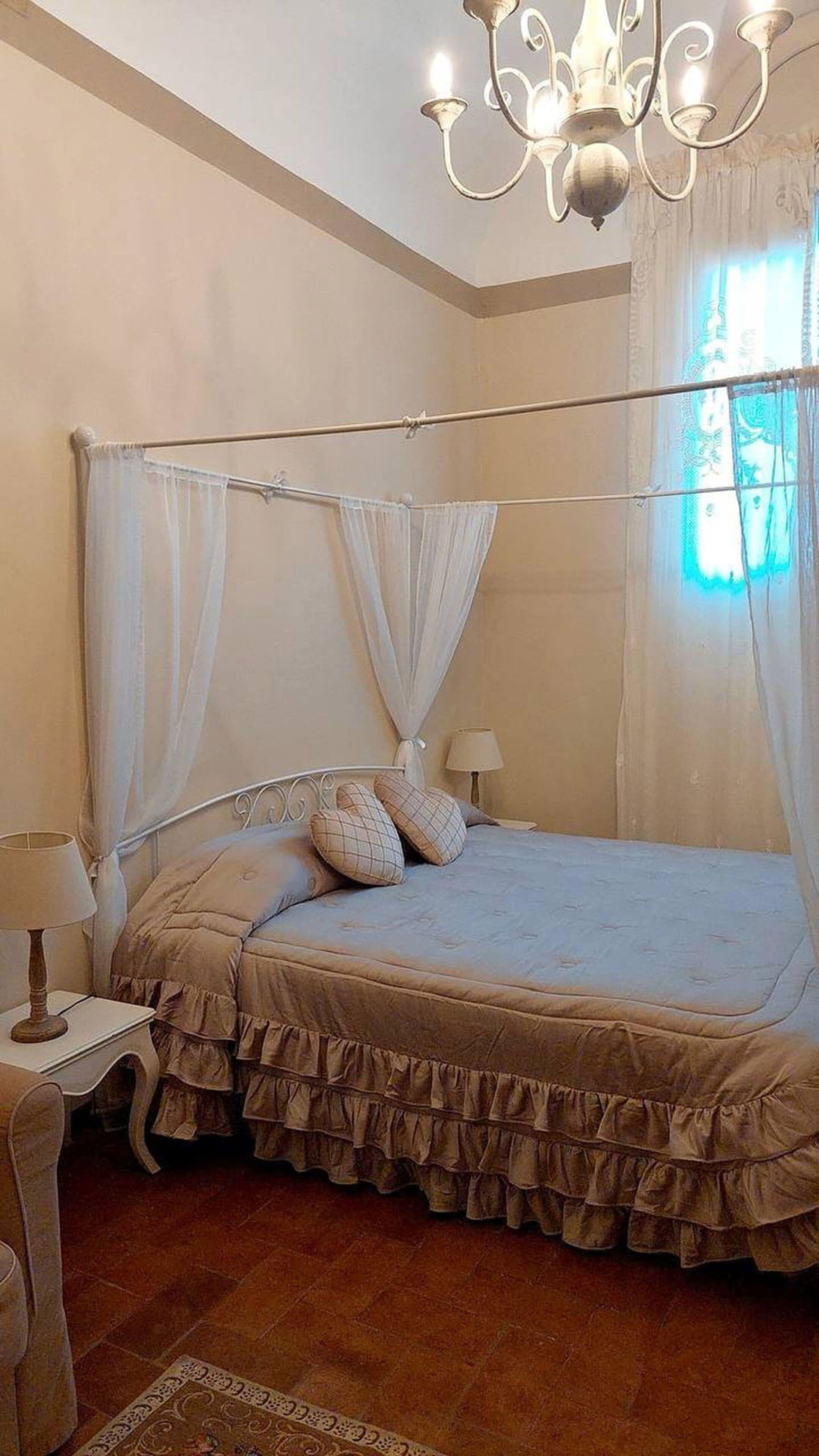 A bedroom with a four-poster bed, a chandelier, a nightstand with a lamp, and a window with curtains.