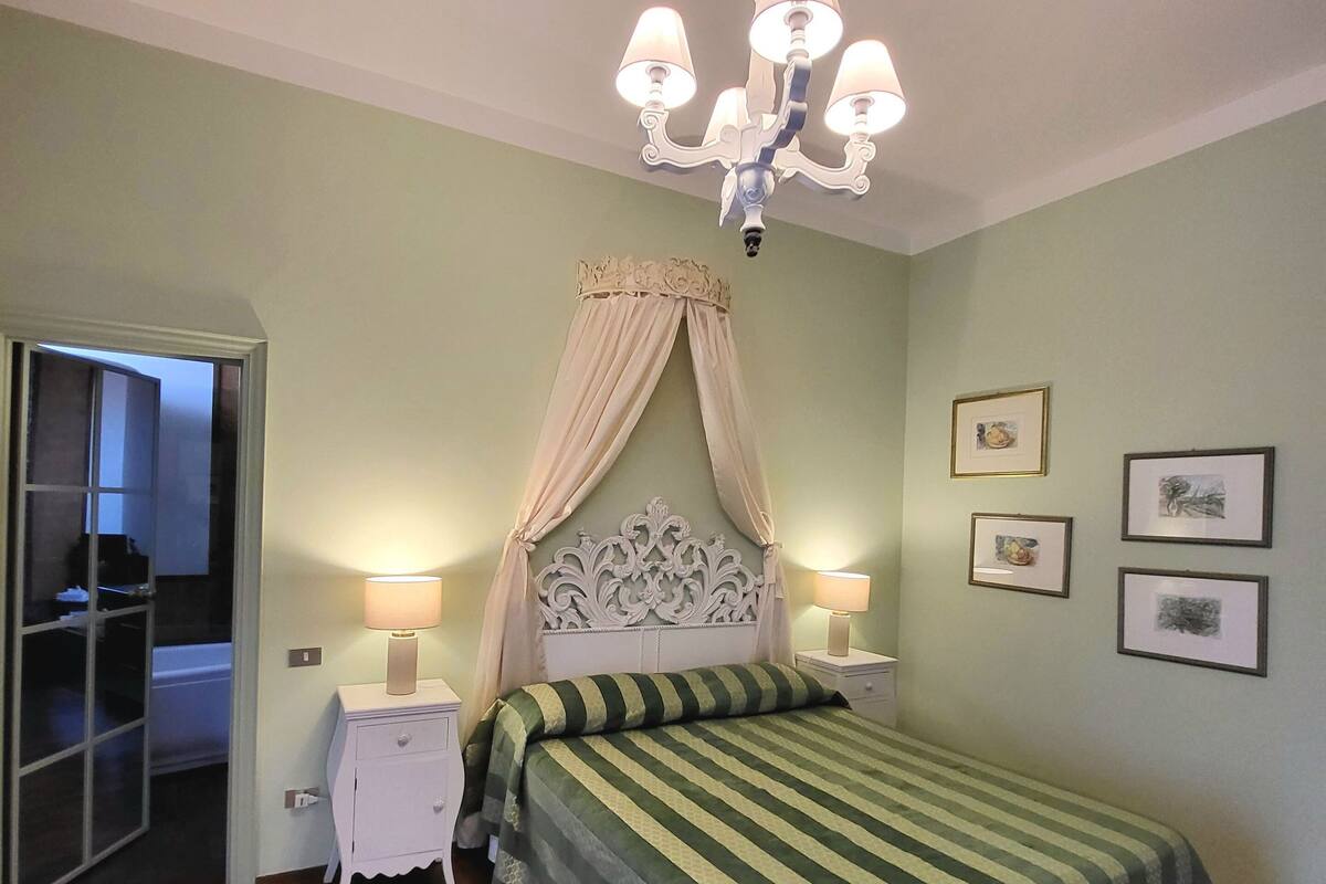 A bedroom with a bed, two bedside tables, a chandelier, and a door leading to another room.