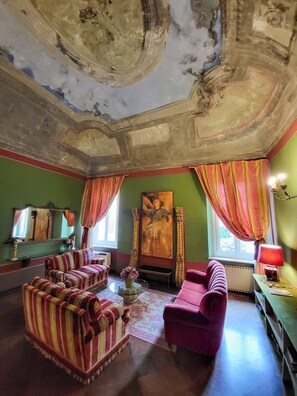 Une pièce avec un plafond haut, des murs verts et un grand tableau. On y trouve deux canapés à rayures et un fauteuil rose.