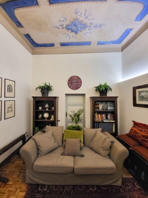 Un salon avec un canapé beige, deux tables d’appoint ornées de plantes, une horloge au mur et une porte surmontée d’un décor.