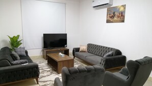 A modern living room with a flat-screen TV, two grey sofas, a wooden coffee table, and a large window. - Balqees Villa (Seeb)