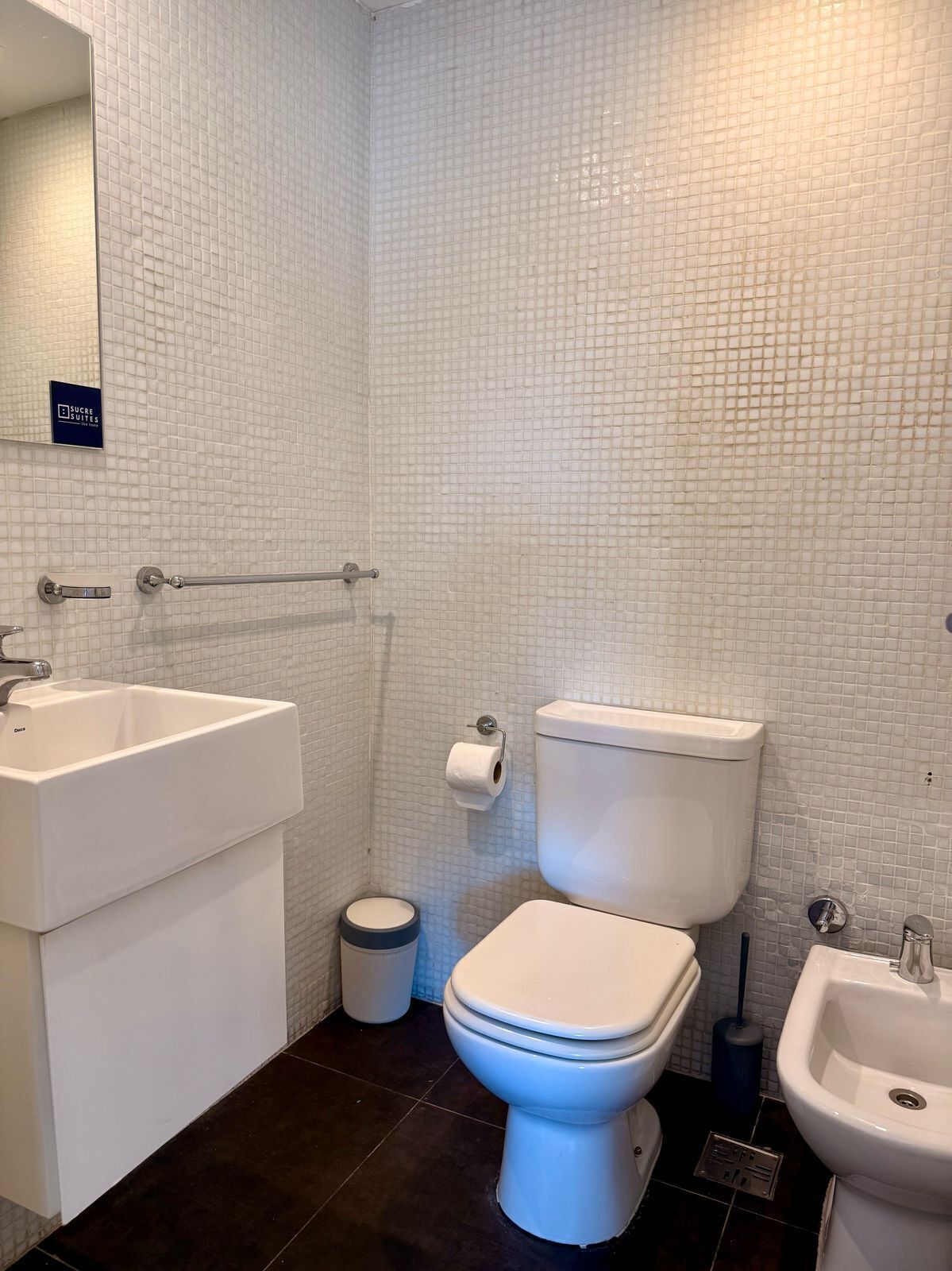 Un baño con pared de azulejos blancos, inodoro, lavabo y cubo de basura.
