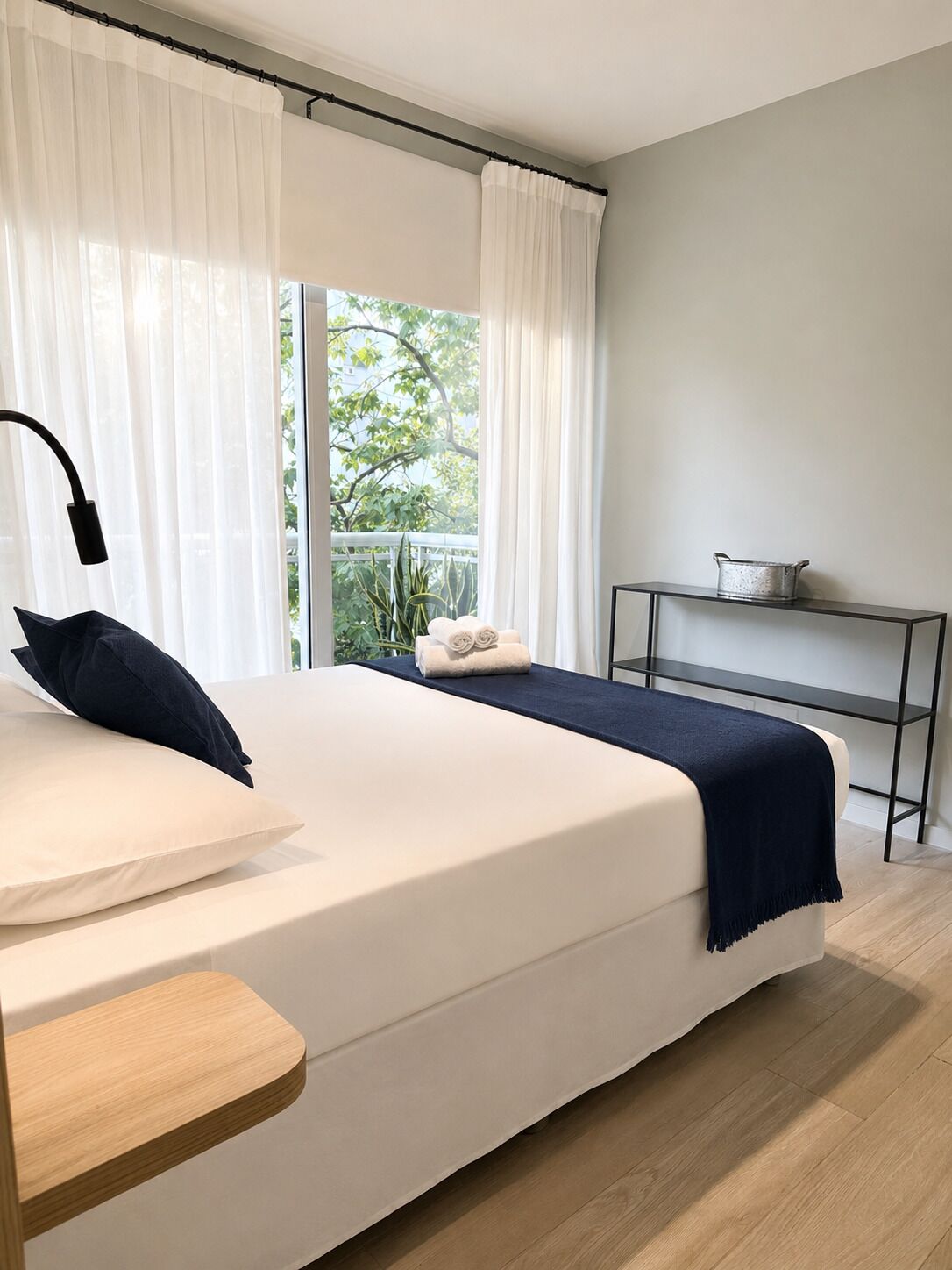 A modern bedroom with a bed, a bedside table, a lamp, a window with curtains, and a small table with a pot.