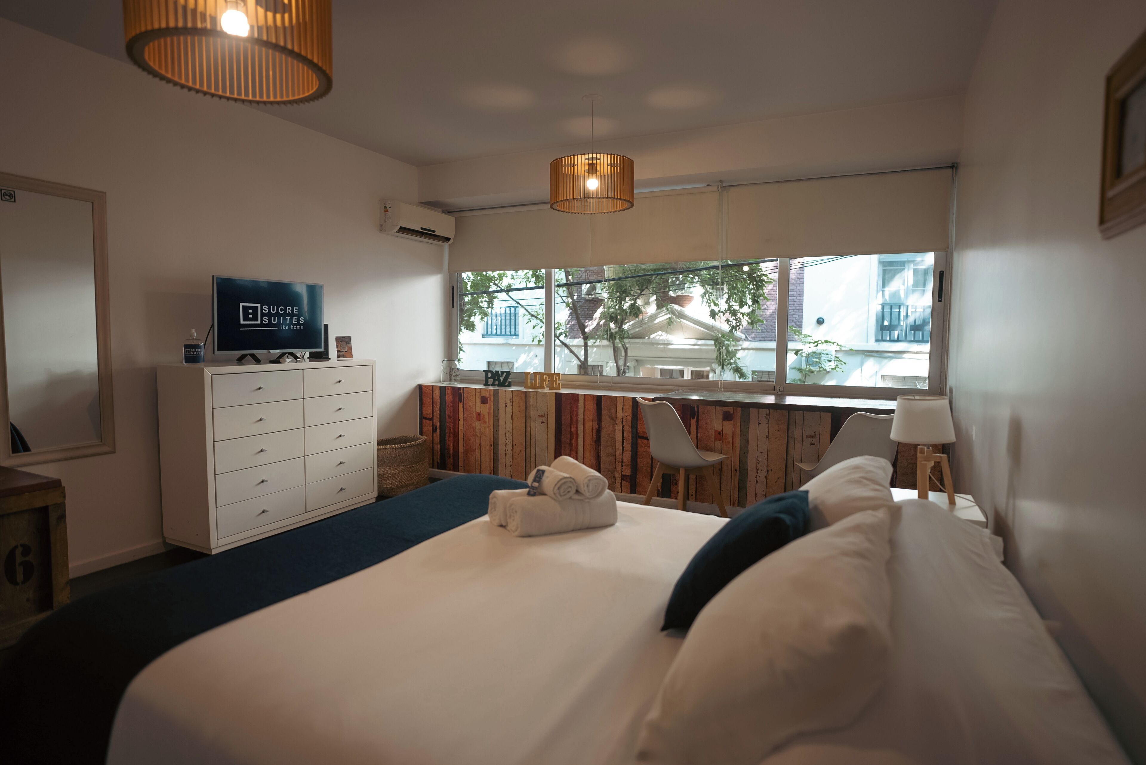 A hotel room with a bed, a dresser, a TV, a window with a view, and a hanging light fixture.
