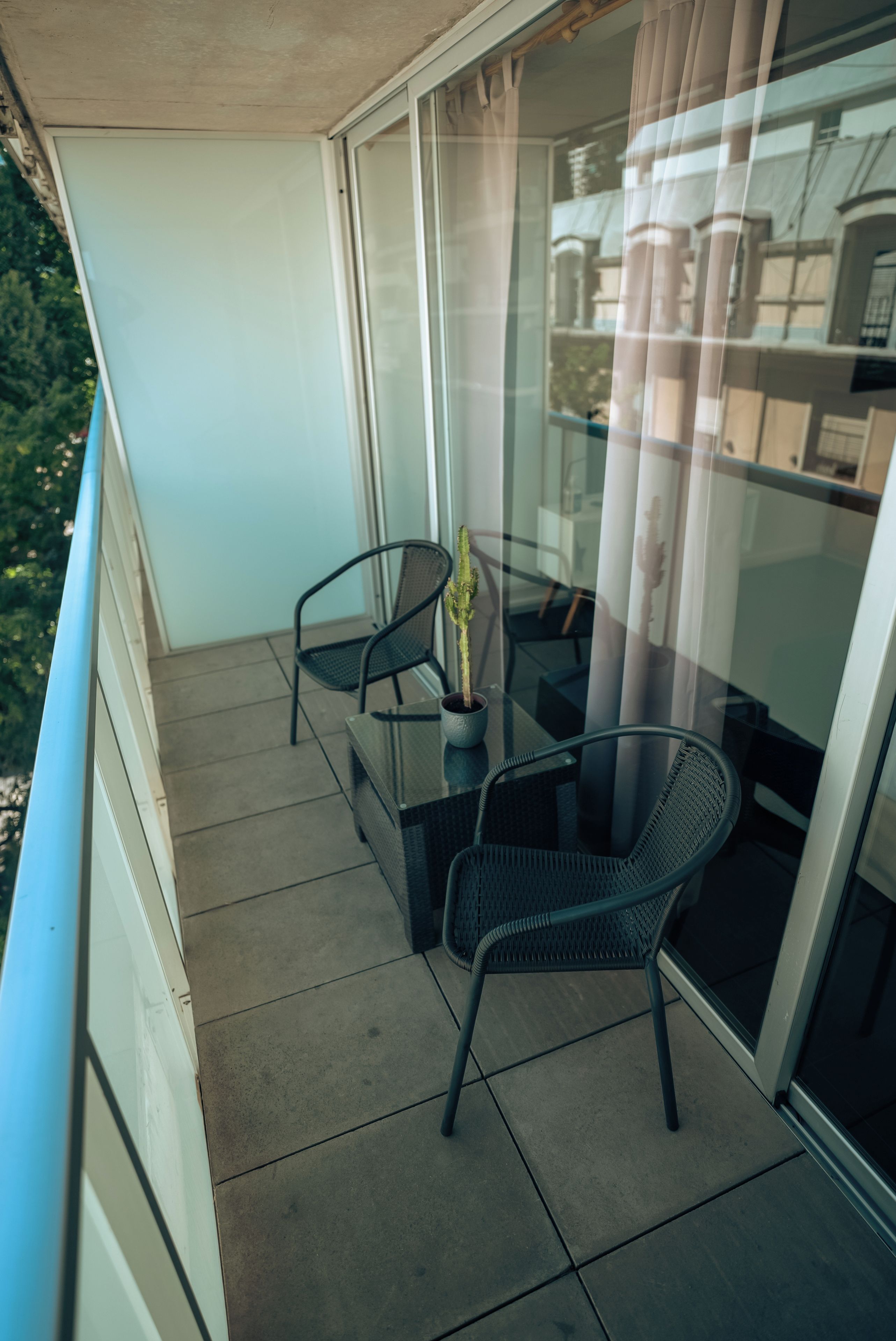 Un balcón con dos sillas, una mesa, una planta en maceta y un ventanal con cortinas.