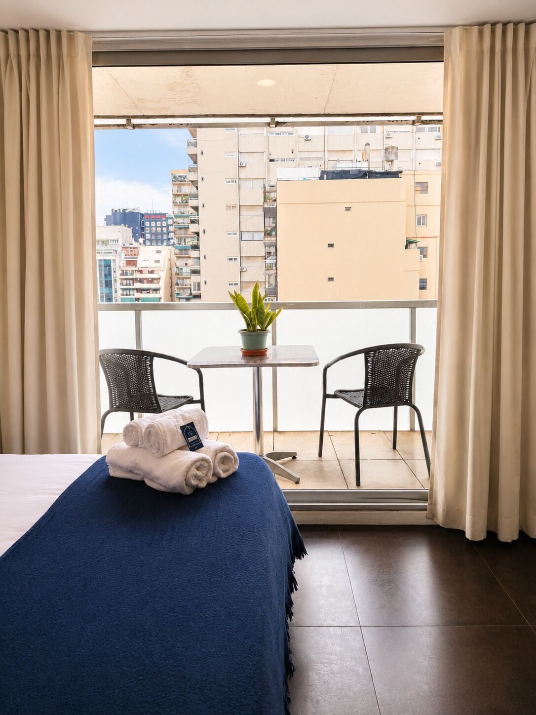 Une chambre d’hôtel avec un lit, une petite table, deux chaises, une plante en pot et une vue sur les bâtiments depuis un balcon.
