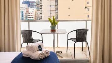 Une chambre d’hôtel avec un lit, une petite table, deux chaises, une plante en pot et une vue sur les bâtiments depuis un balcon.