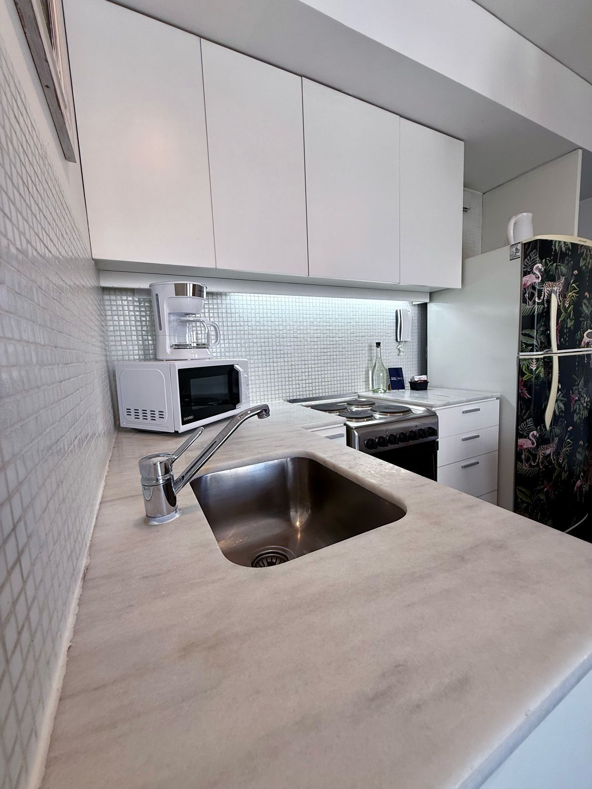 Uma cozinha moderna com armários brancos, pia de aço inoxidável, micro-ondas e geladeira com design tropical.