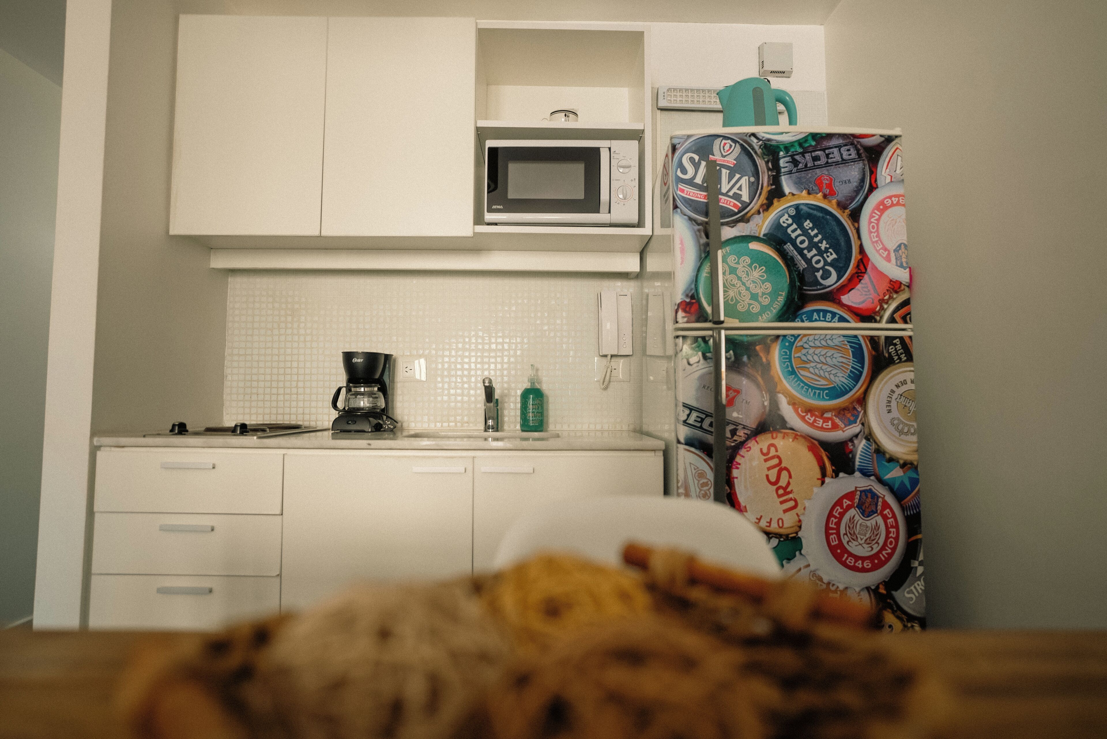 Una cocina con armarios blancos, microondas, cafetera y una estantería con tapones de botellas de cerveza.